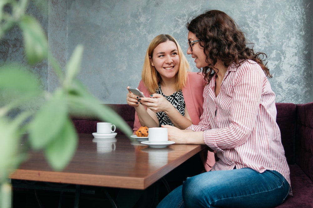 Foto van twee vrouwen aan tafel die op mobiel kijken